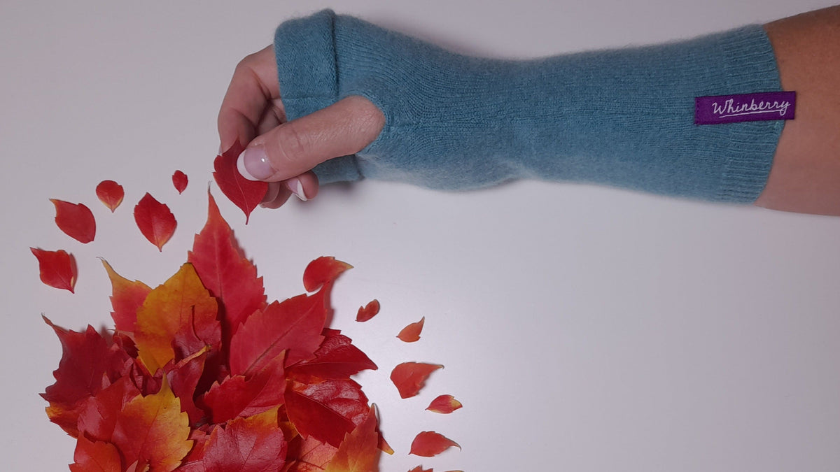 A person's arm wearing a sky blue whinberry fingerless glove picking up an autumnal red leaf from a spray of leaves